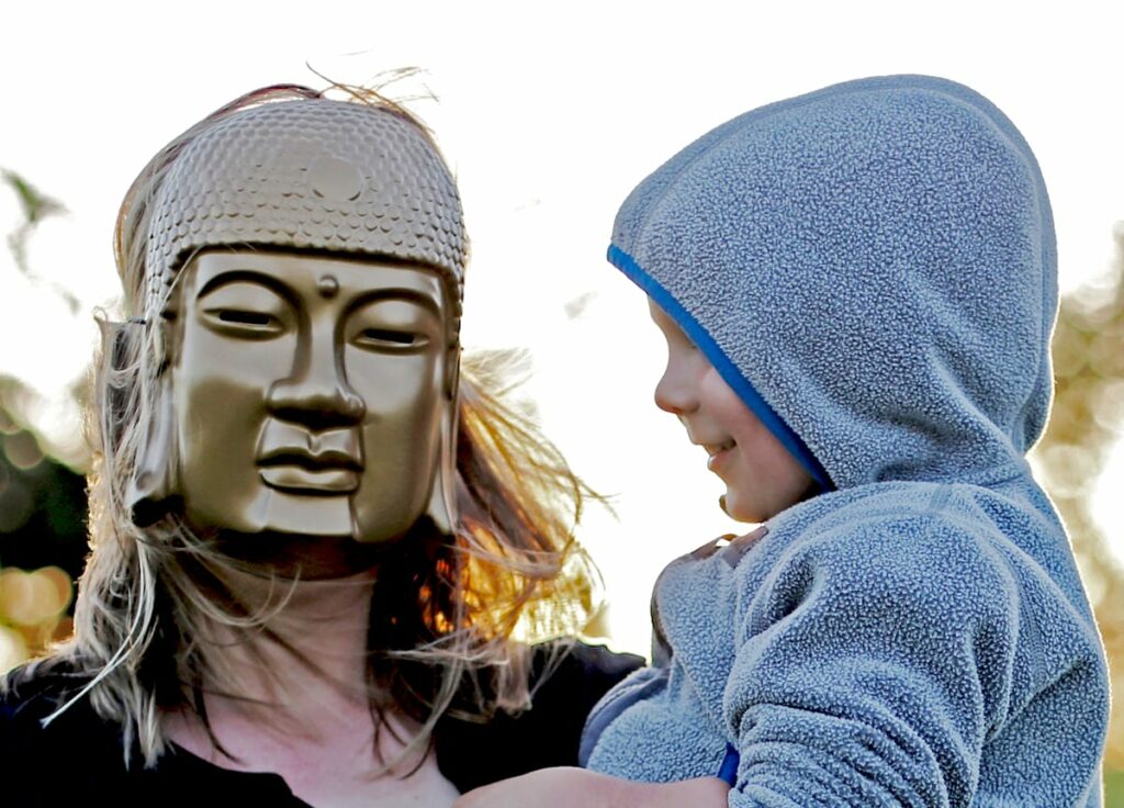 Mother wearing Buddha mask holding lughing child.
